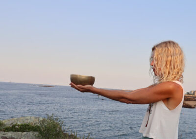 A Profile View Of Trish Hart, Arms Extended Holding A Bowl With The Ocean In The Background